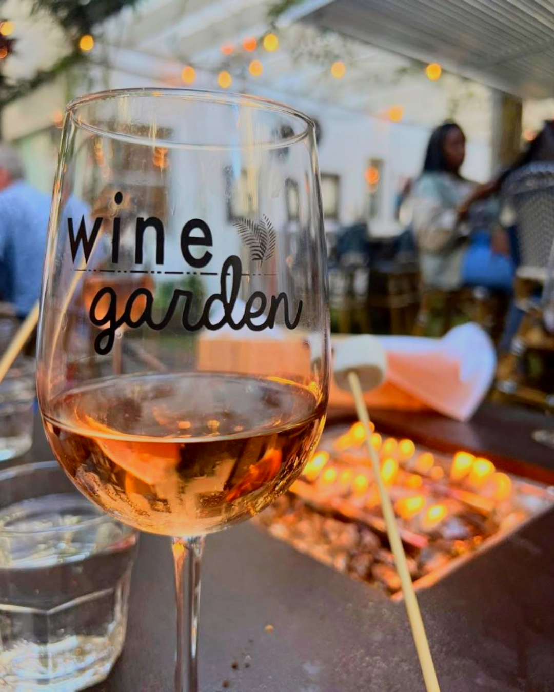 A wine glass with 'wine garden' written on it, sitting on a table with a fire pit in the background.