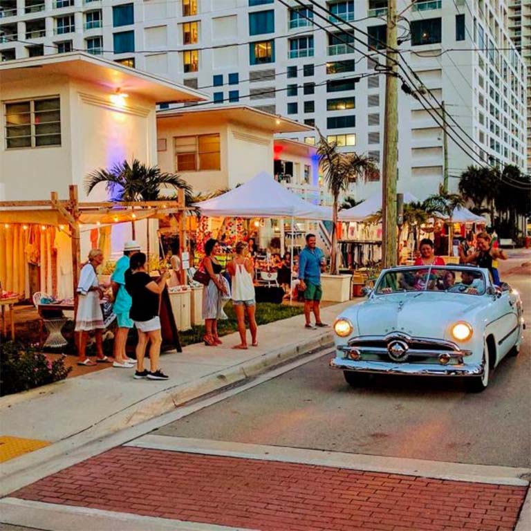 A white car drives down a street with people gathered outside a building with tents and palm trees.